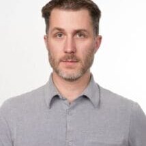 Portrait of a man with short brown hair and light stubble wearing a gray shirt.