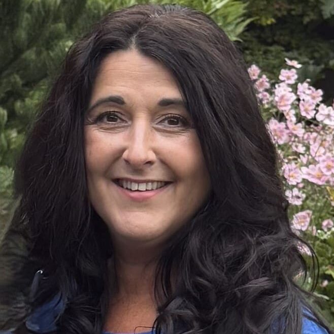 Smiling woman with dark curly hair outdoors near flowering tree.